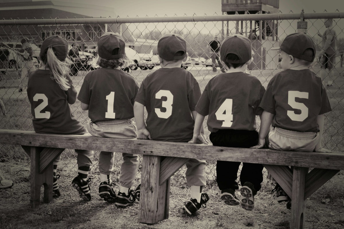 Kids in numbered baseball jerseys on a bench, seen from behind
